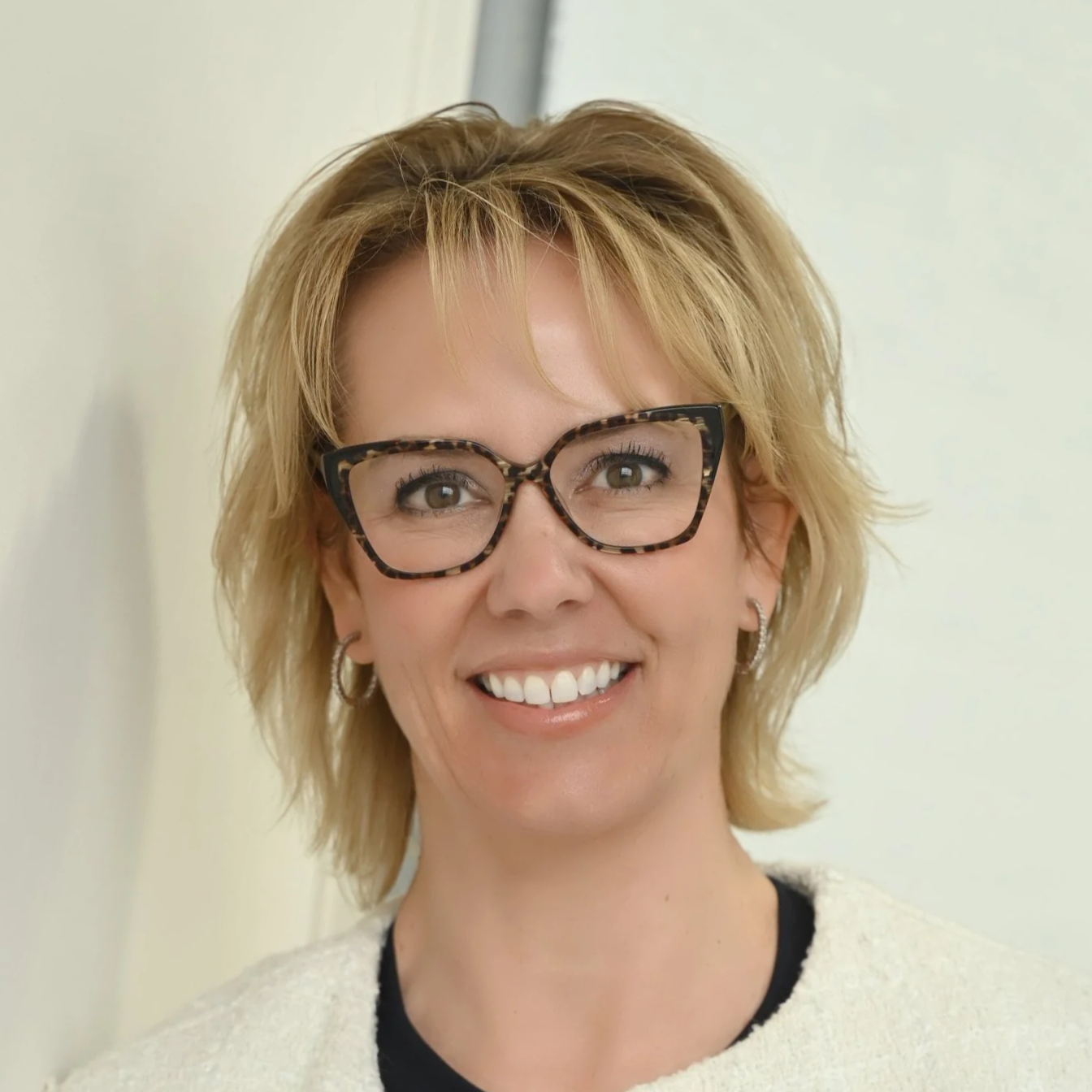 Woman with blonde hair, wearing black-rimmed glasses, smiling, against a neutral background.