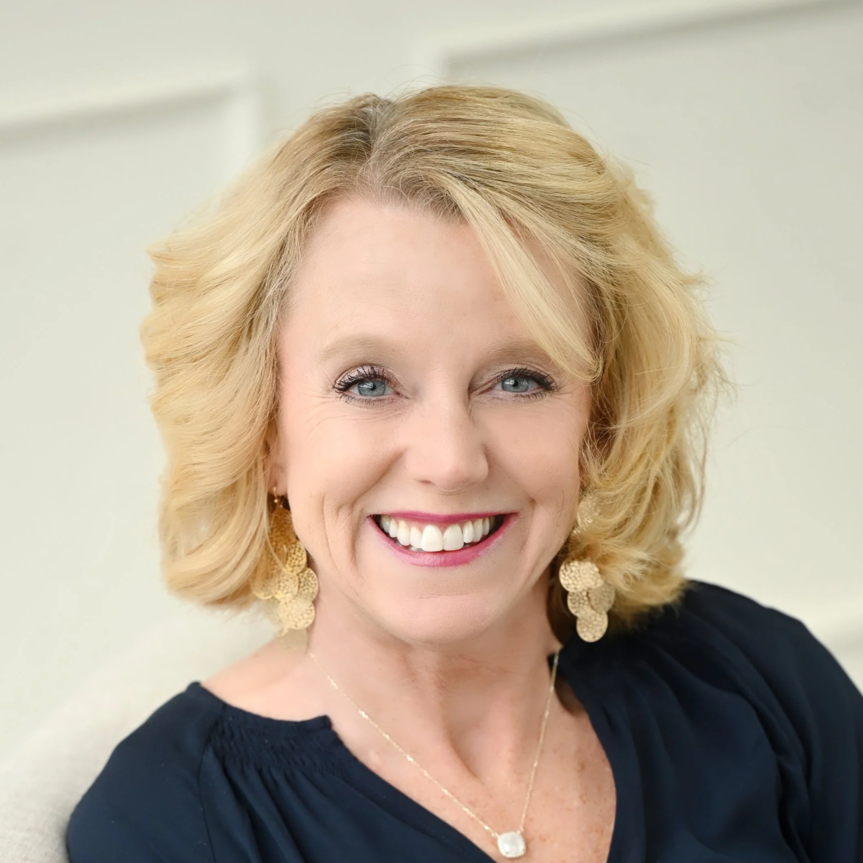A woman with blonde, shoulder-length hair, smiling, wearing a black top, gold earrings, and a gold necklace.
