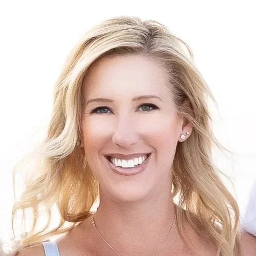 A woman with blonde hair smiling at the camera, wearing a light-colored top and small earrings.