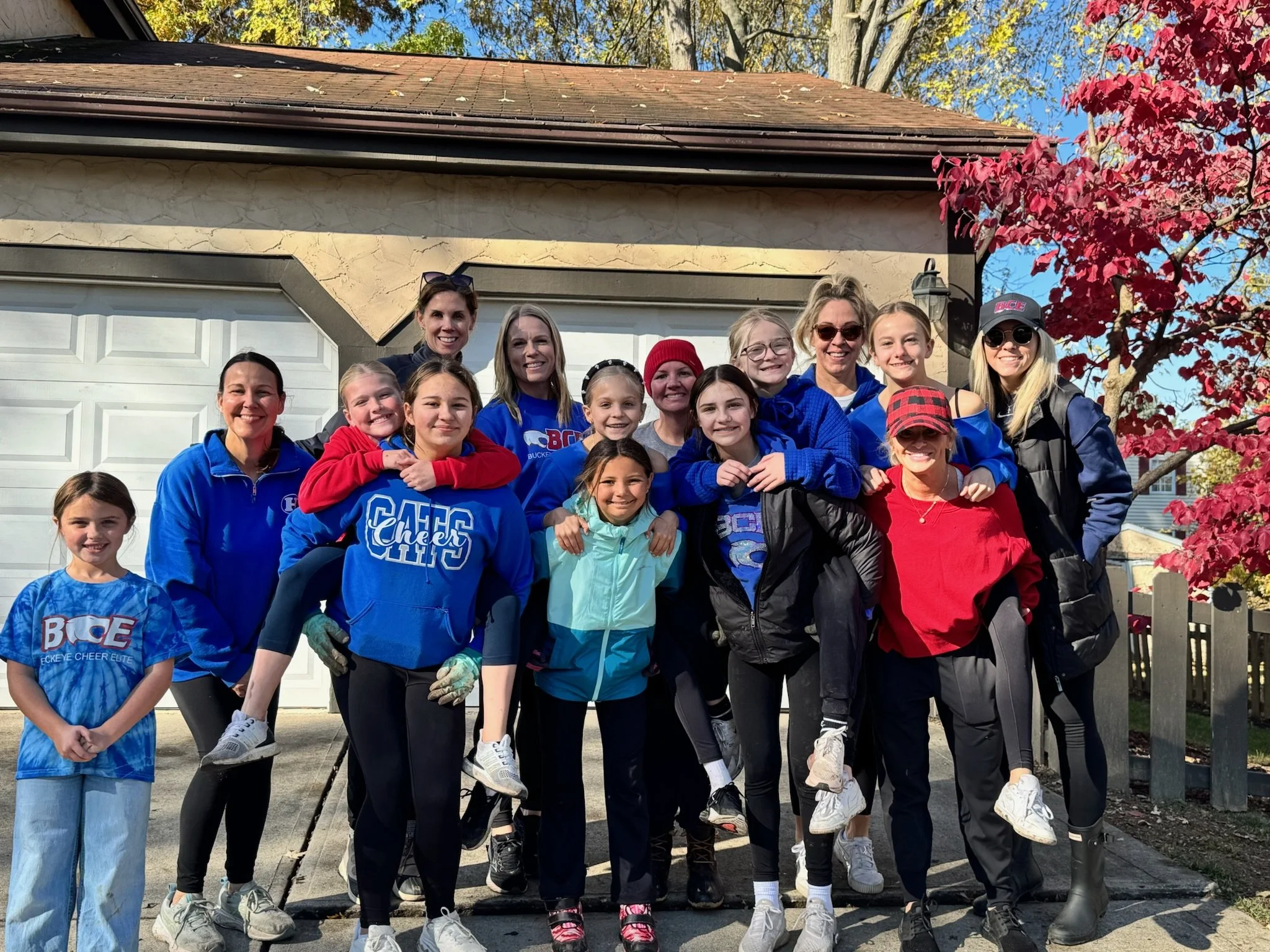 A group of smiling women and children, some with their arms around each other, pose together outside a house with a garage door, red and yellow autumn leaves on a tree, and a blue sky in the background.