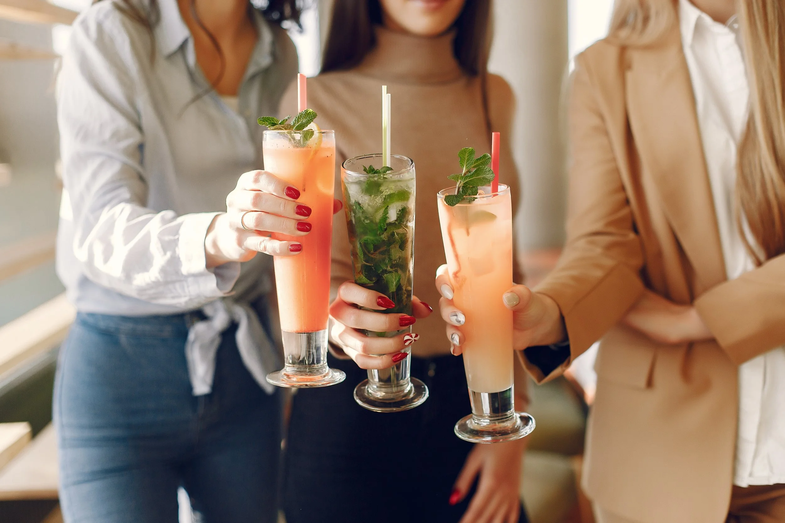Three women holding colorful cocktails with straws and mint garnishes