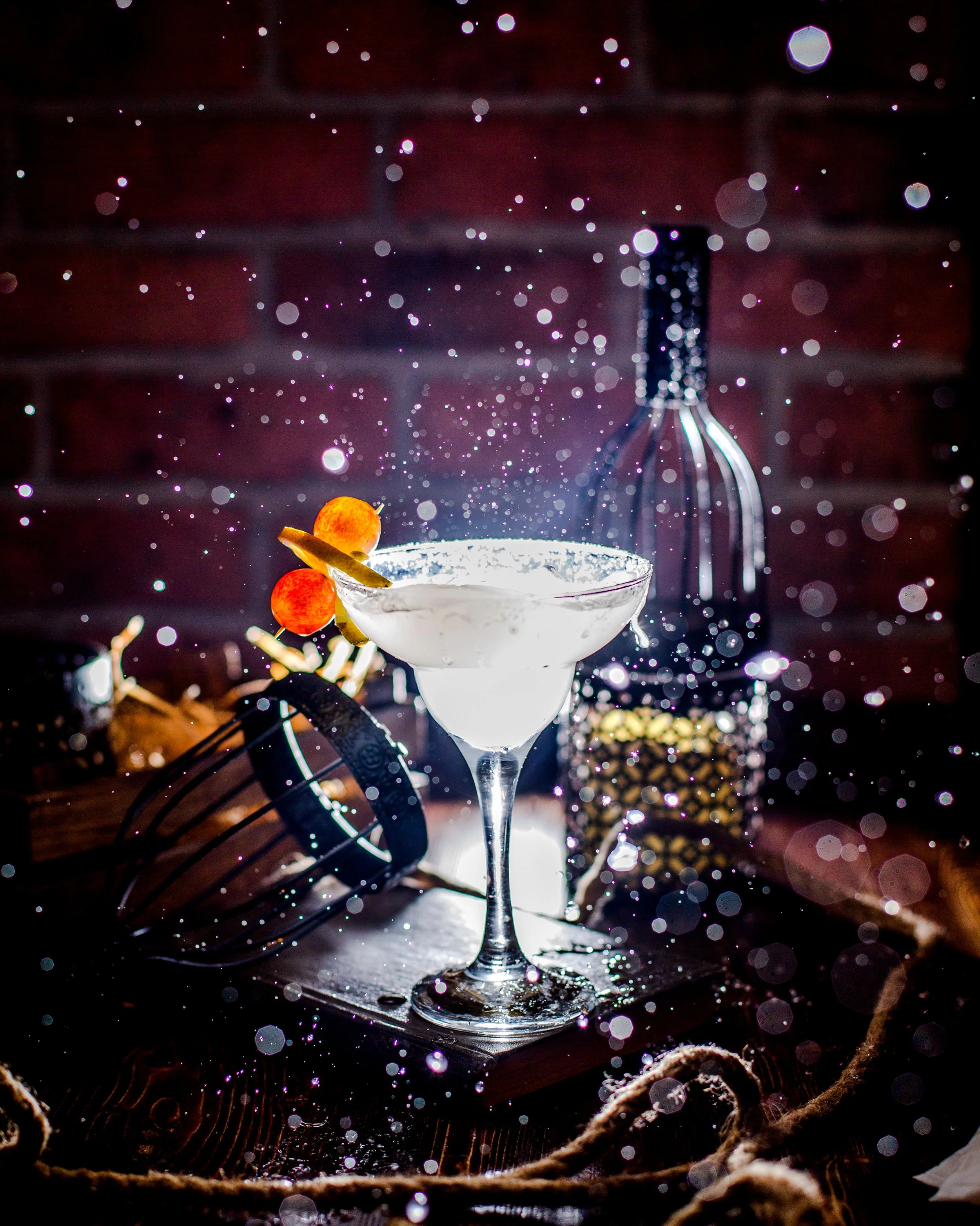 A cocktail glass with a white drink, garnished with red berries and a lemon peel, on a wooden surface with a blurred background, surrounded by bokeh lights, a whisk, and spices.