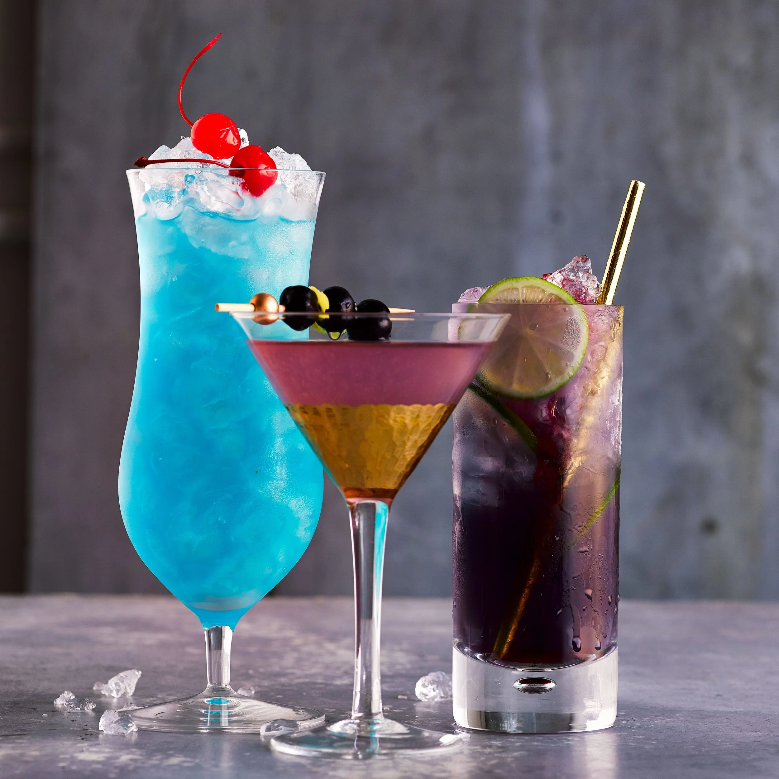 Advert for our cocktail making class - Three colorful cocktails on a gray surface with ice cubes, with a gray background.