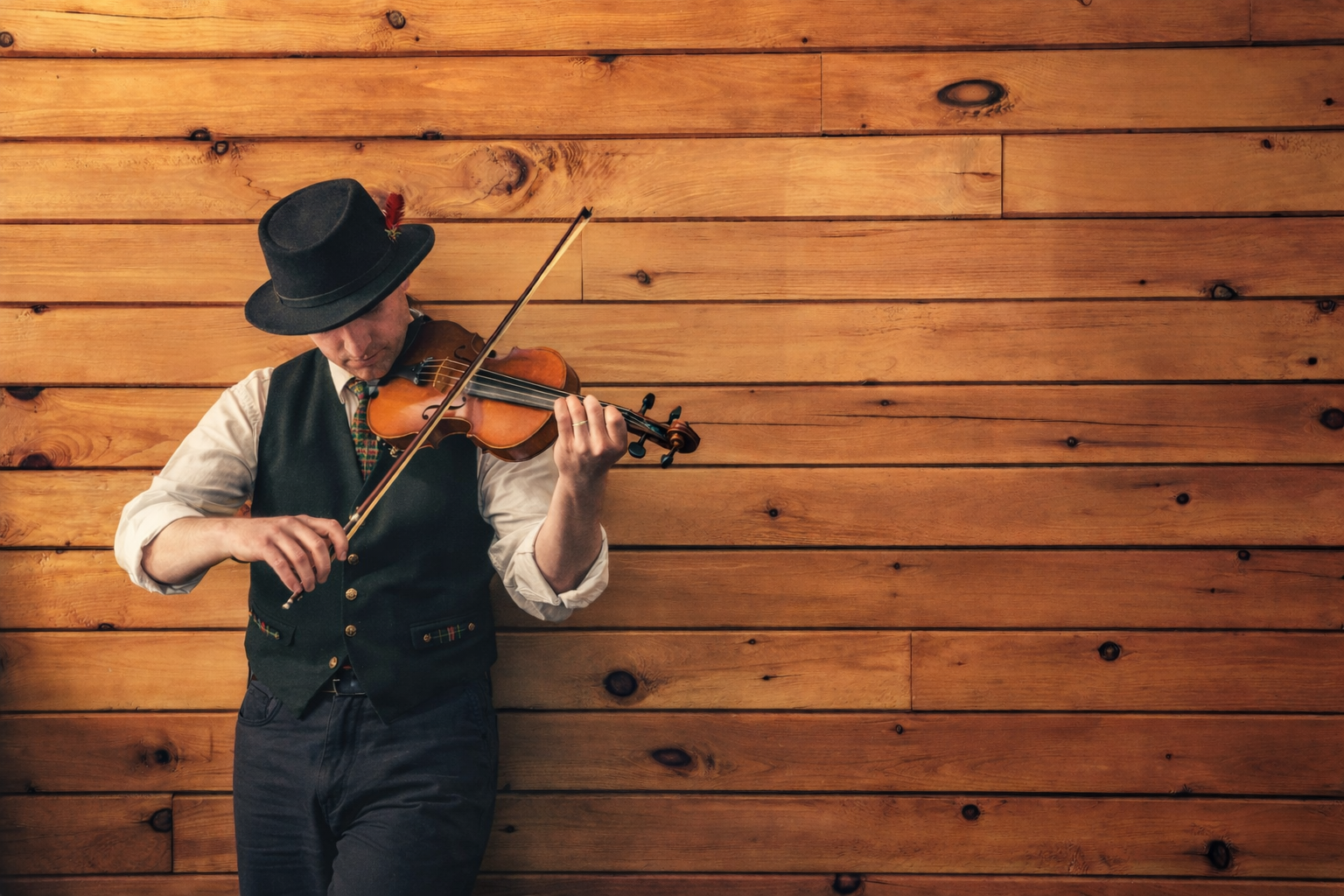 Advert for our Charity Ceilidh - A man wearing a black hat, white shirt, vest, and dark pants playing the violin against a wooden wall.