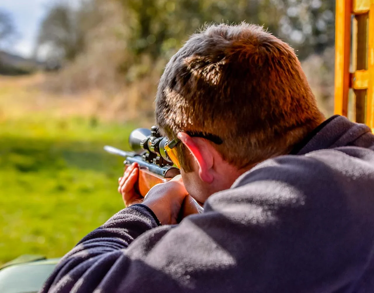 Advert for our air rifle competition - A man with short brown hair and sunglasses aiming a rifle with a scope outdoors, with a blurred green natural background.