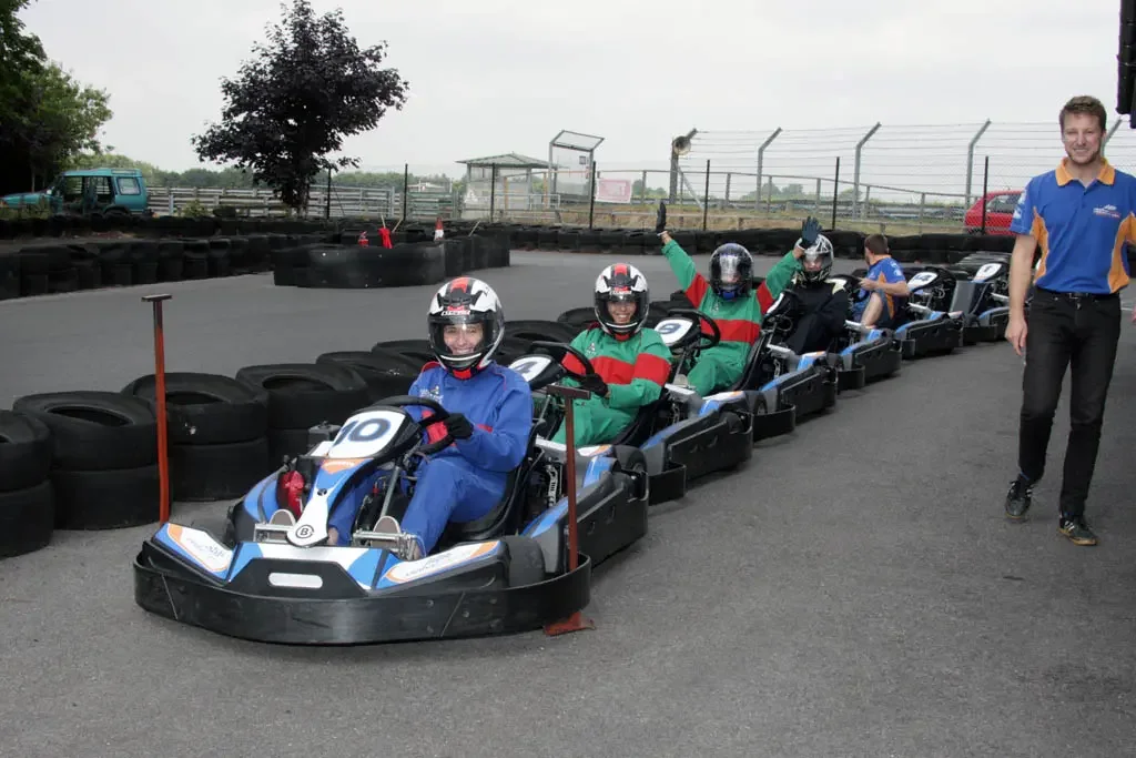 Advert for our go-karting night - Three people in go-karts on a race track, wearing helmets and racing suits, with a staff member standing nearby.