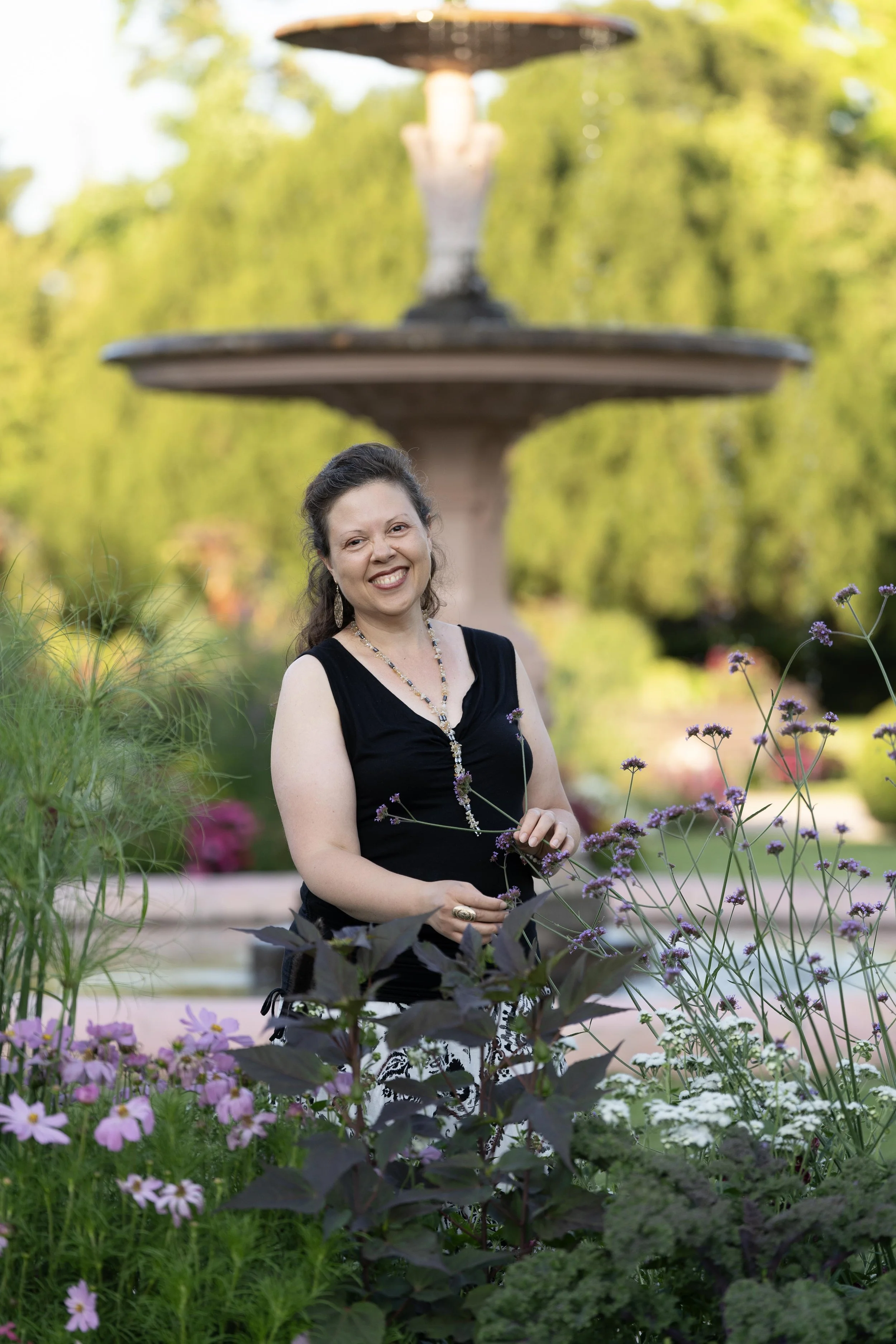 Une femme souriante en robe noire posant dans un jardin fleuri avec une fontaine en arrière-plan.