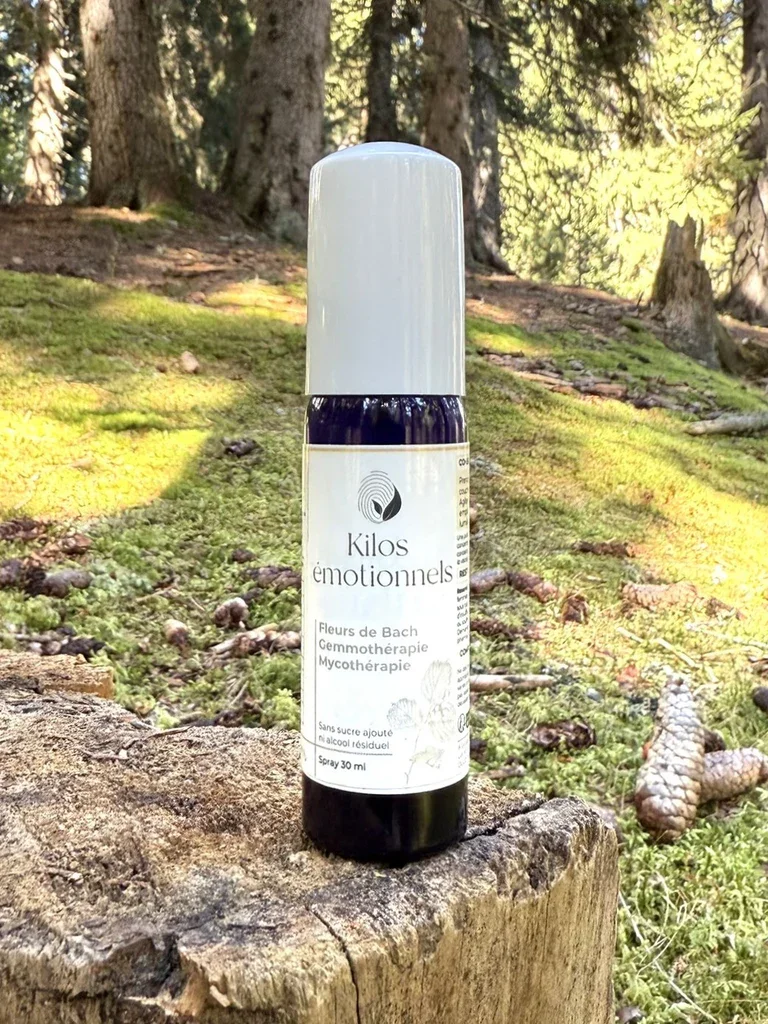 Un flacon de spray sur un tronc d'arbre en forêt, label indiquant 'Kilos émotionnels, Fleurs de Bach, Gemmothérapie, Mycothérapie', volume 30 ml.