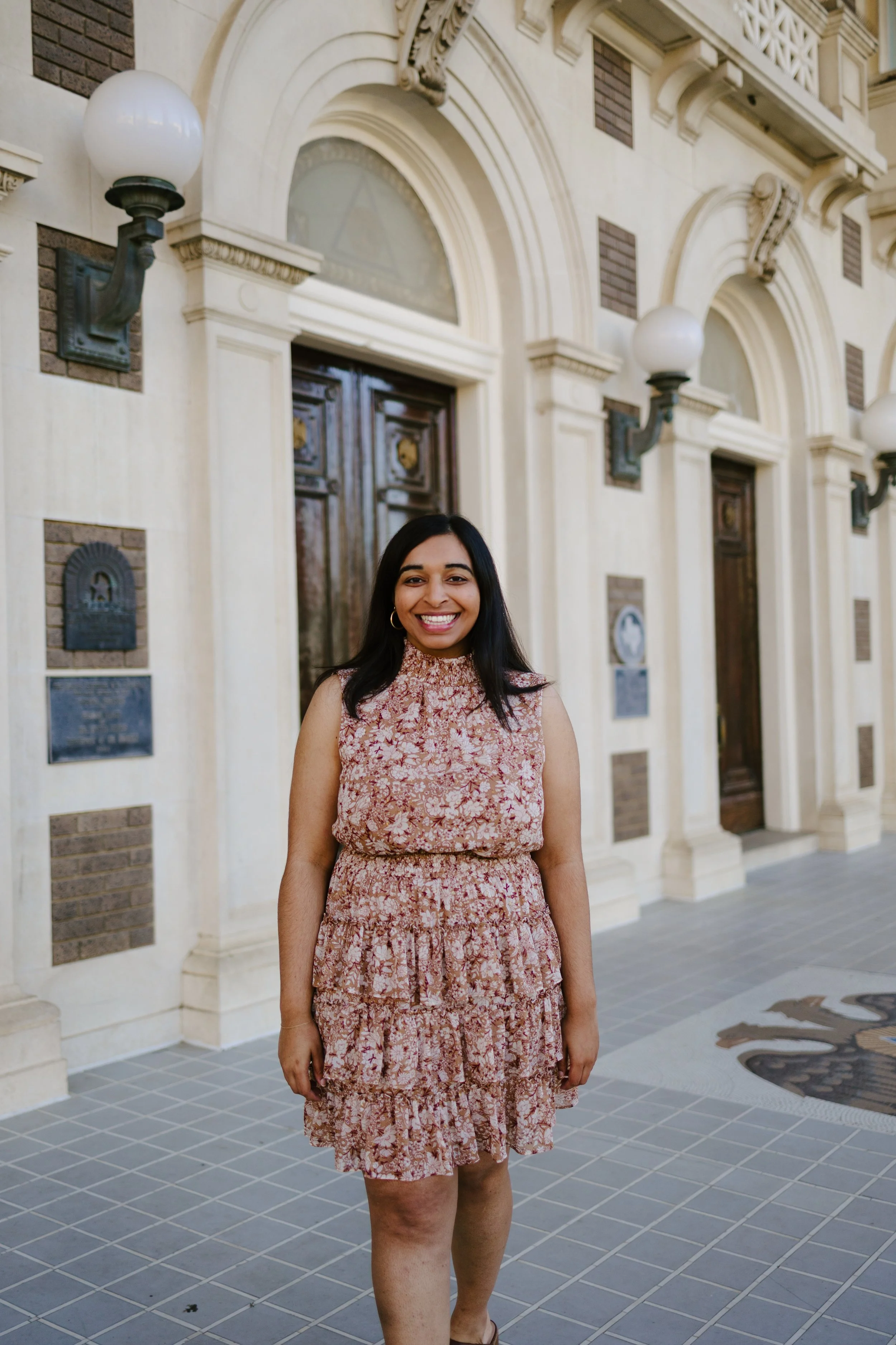 A woman in a pink floral dress standing outside in front of a historic building with arched windows and decorative plaques.