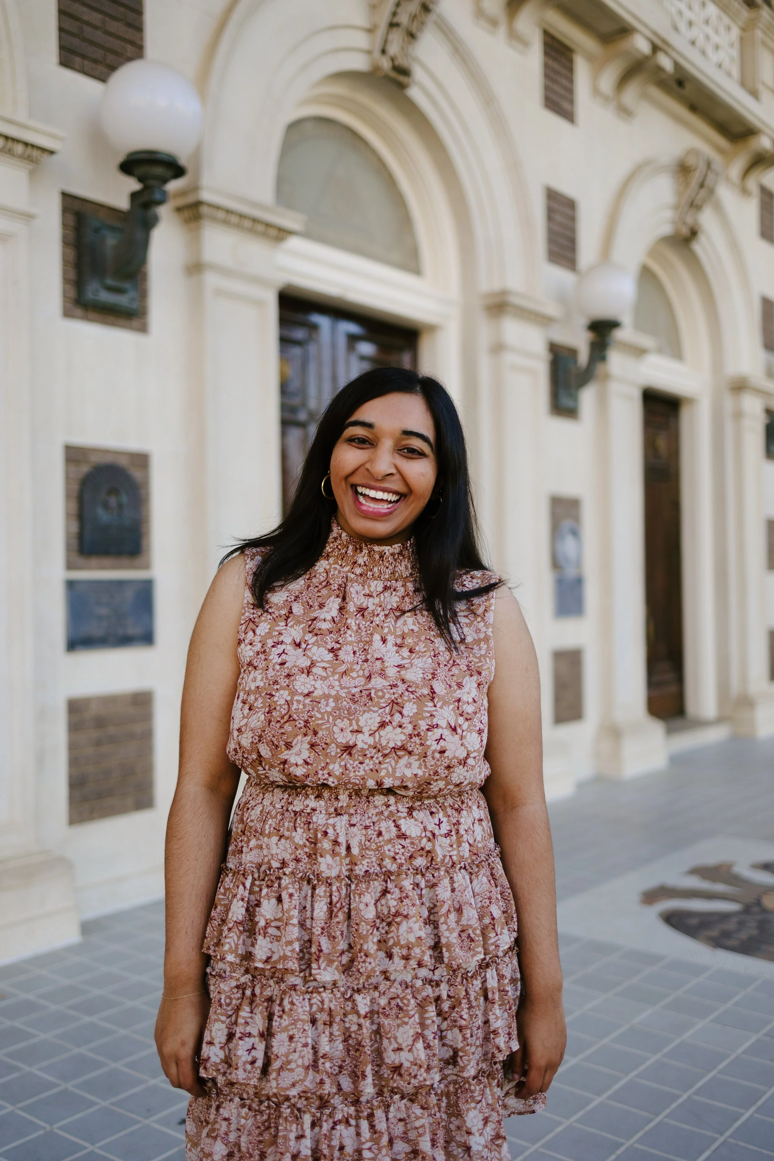 A woman with dark hair, wearing a pink floral dress, smiling and standing in front of an ornate building with arched windows and wall-mounted lamps.