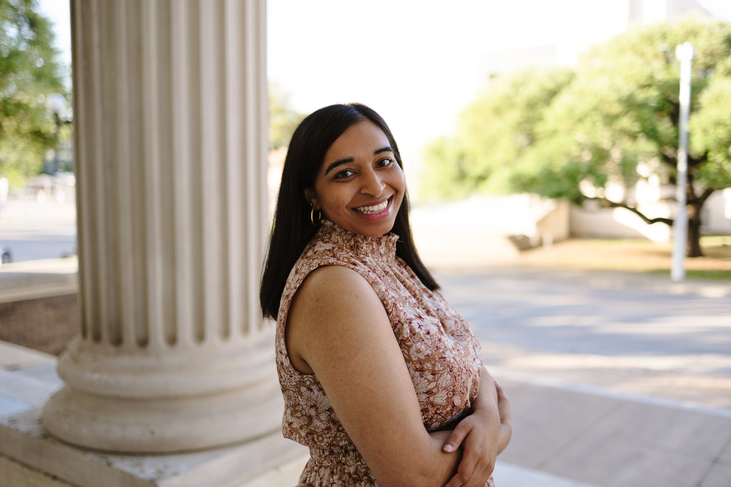 A woman with dark hair smiling outdoors next to large columns on a sunny day.