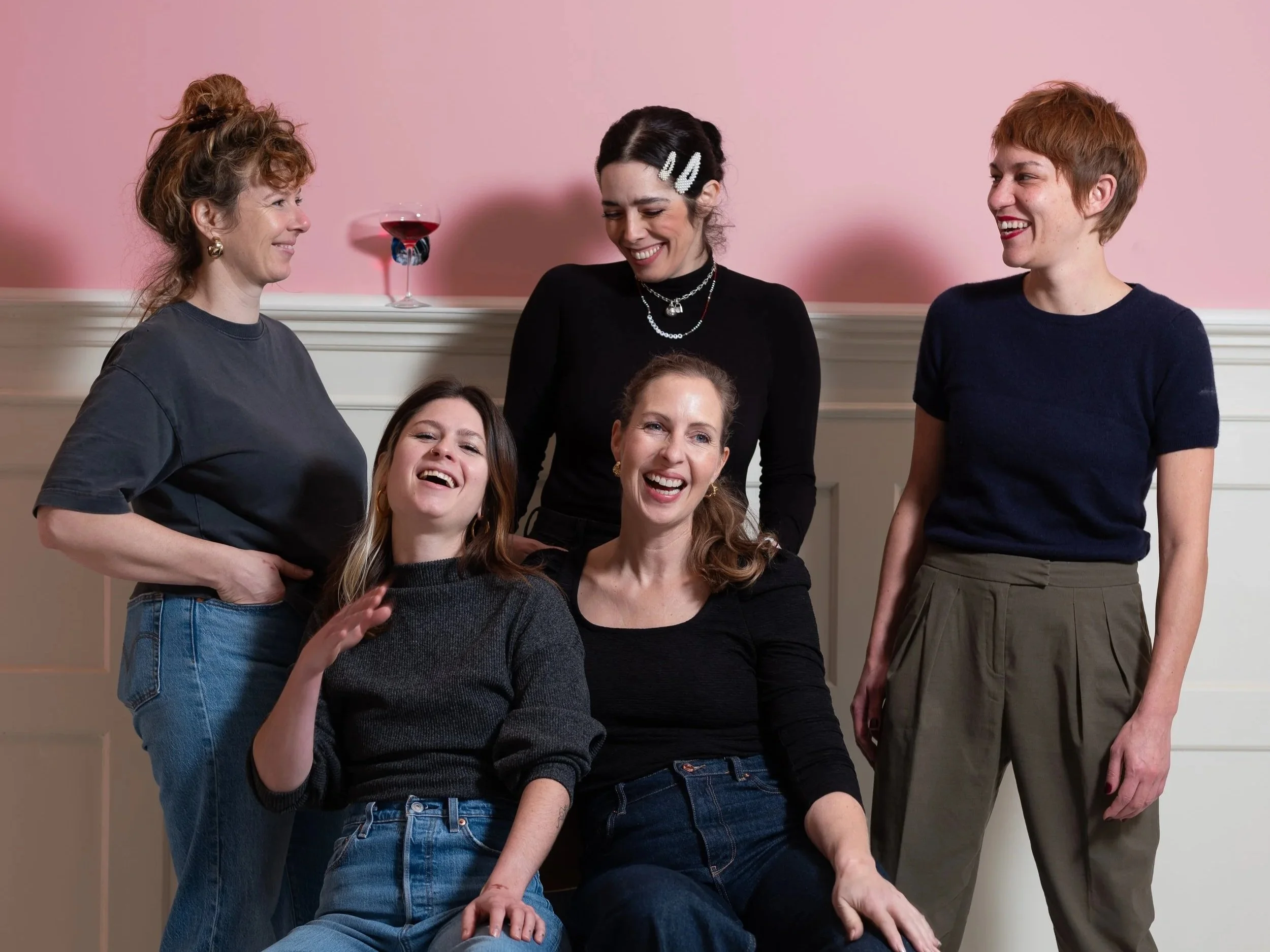 Sechs Frauen lachen und lachen bei einer Party in einem Raum mit rosa Wand und Wein. Einige sitzen, andere stehen und alle scheinen Spaß zu haben.