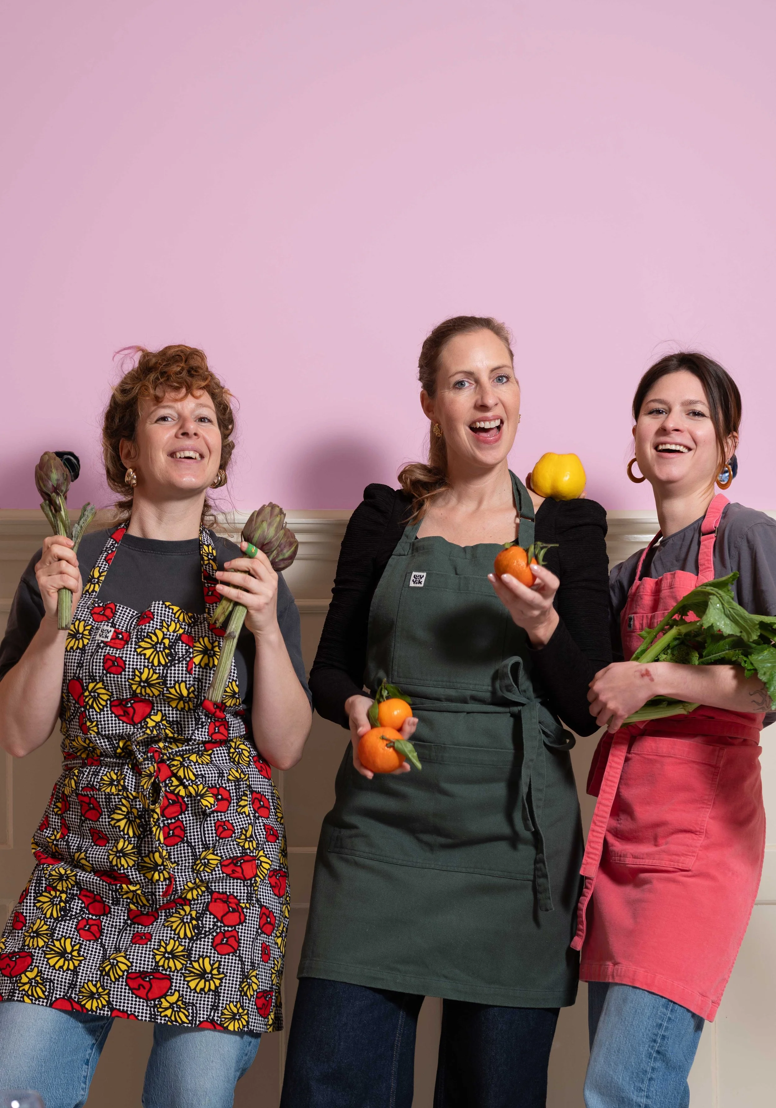 Drei Frauen tragen bunte Schürzen und halten frisches Gemüse, während sie vor einer rosa Wand lächeln.