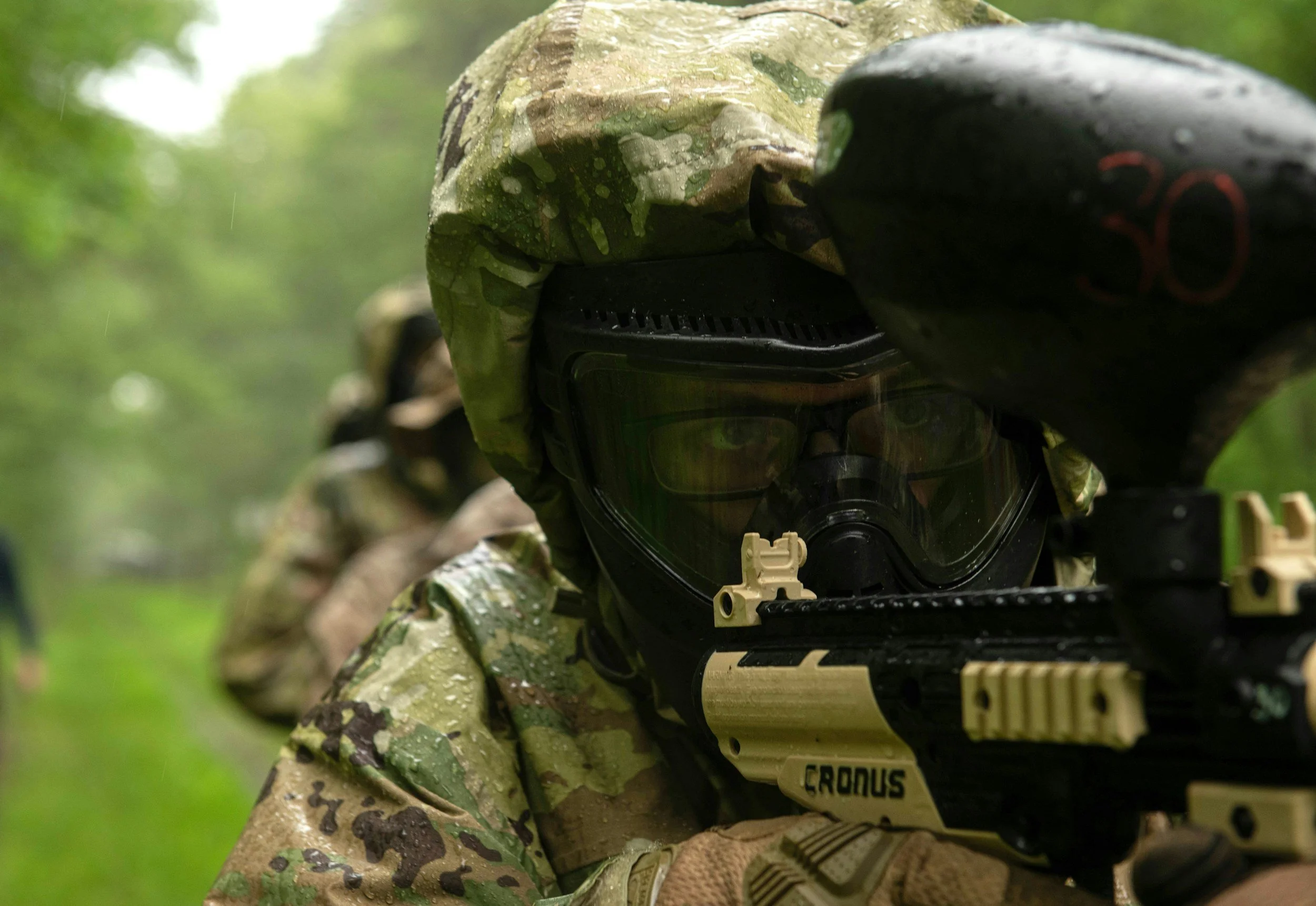 Soldado em equipamento de camuflagem durante treino com arma, usando máscara de proteção facial e óculos, em ambiente ao ar livre com árvores ao fundo.