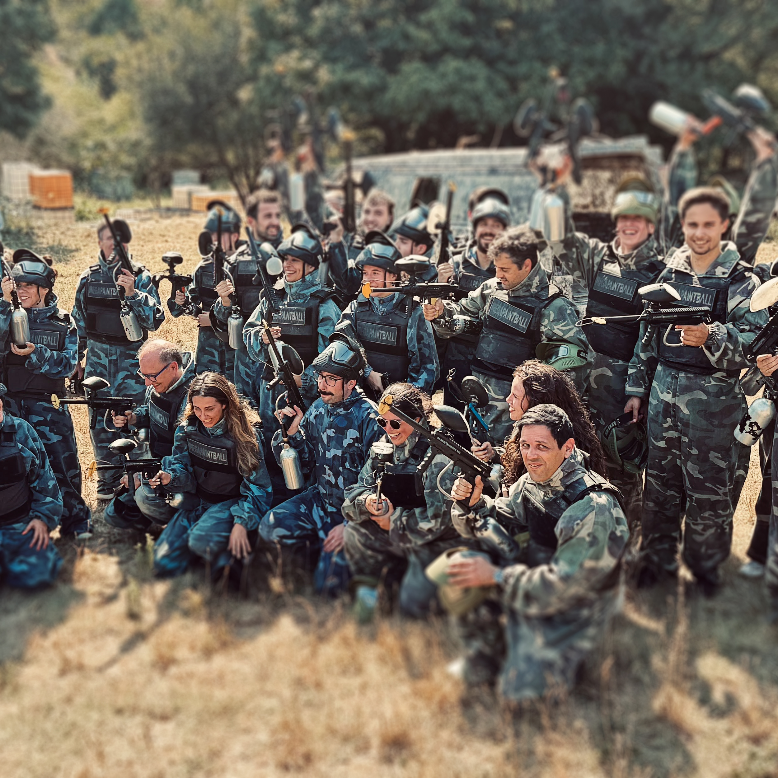 Grupo de pessoas usando roupas de combate e equipamentos de paintball, segurando pistolas de paintball, em um campo ao ar livre.