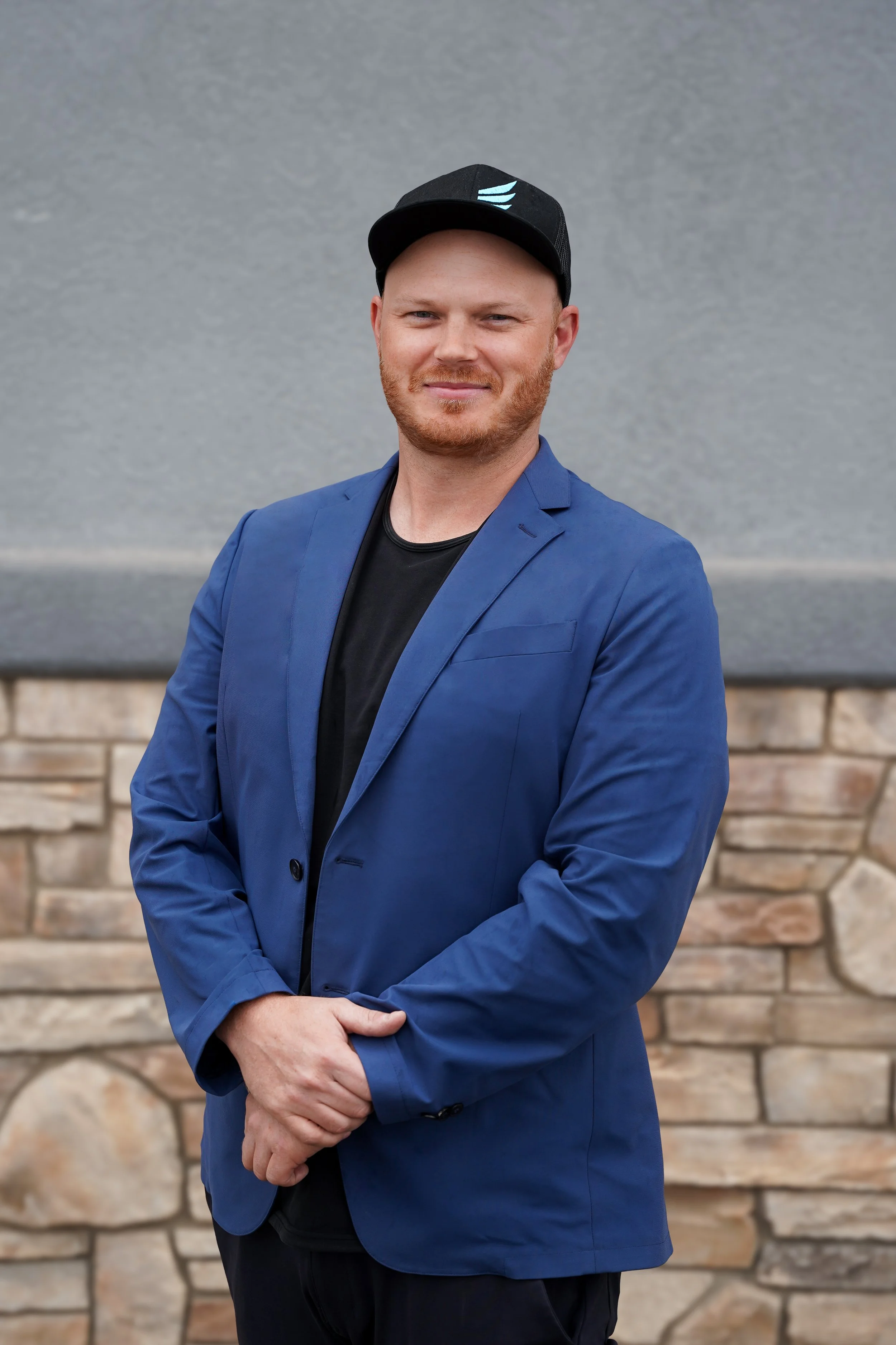 A man with red hair and a beard wearing a black baseball cap, a blue blazer, and a black T-shirt, standing outdoors in front of a gray wall and stone wall.
