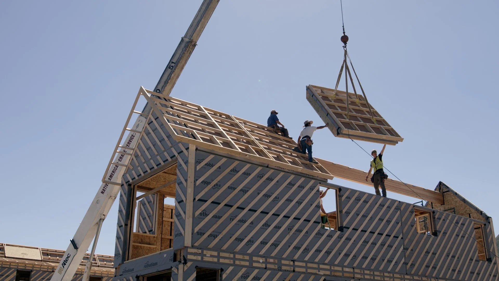 Construction workers installing a roof on a multi-story building with a crane lifting a roof panel.