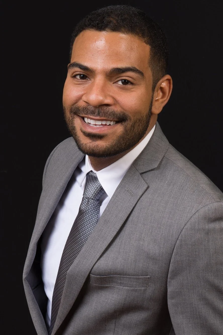 A smiling man in a gray suit, white shirt, and patterned gray tie, posed against a black background.