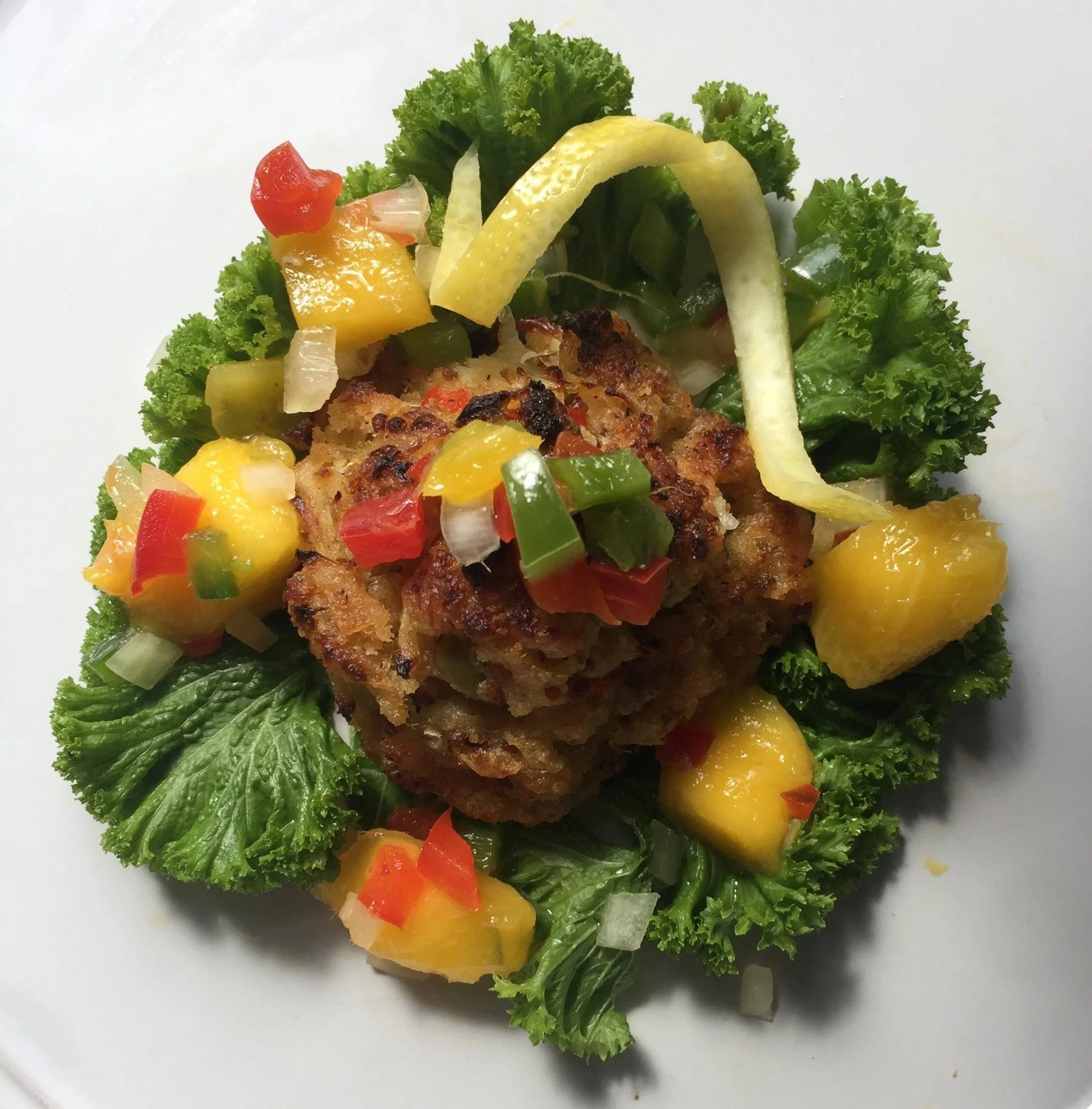 Vegetarian dish with a large, crispy veggie patty, surrounded by lettuce, diced pineapple, chopped red and green peppers, onions, and lemon zest on a white plate.