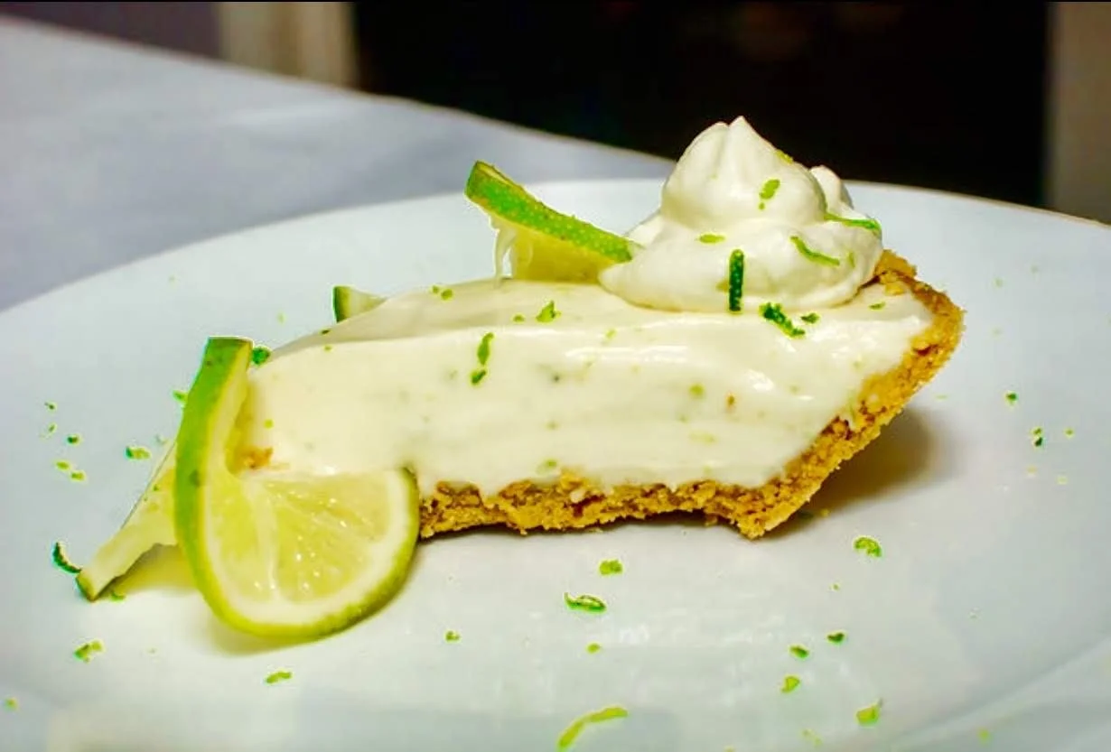 Mango key lime pie with lime slices and whipped cream on top, garnished with lime zest and a lime wedge on a white plate.