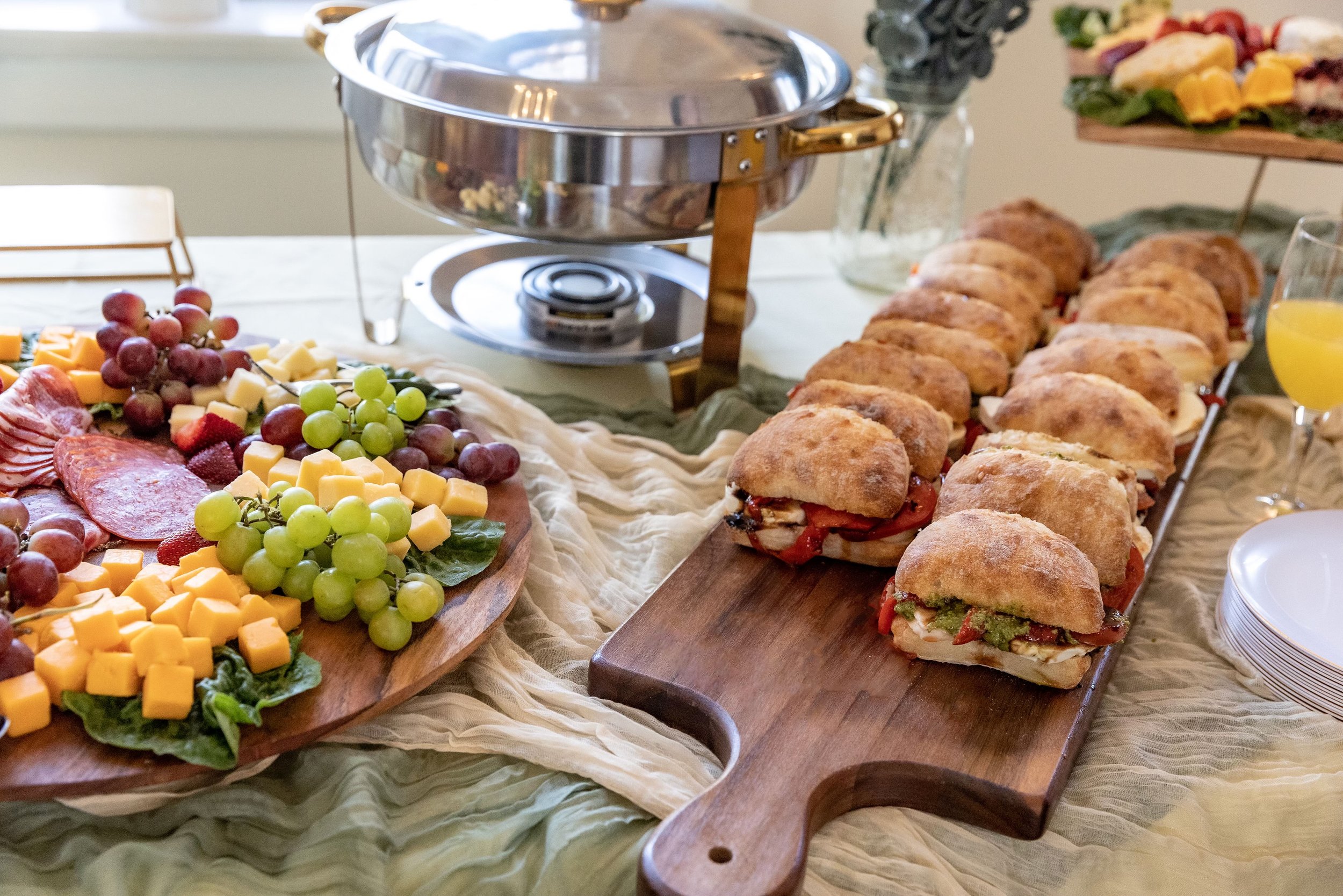 Assorted sandwiches on a wooden platter next to a cheese and grape platter, with a chafing dish and glasses of juice on a table.