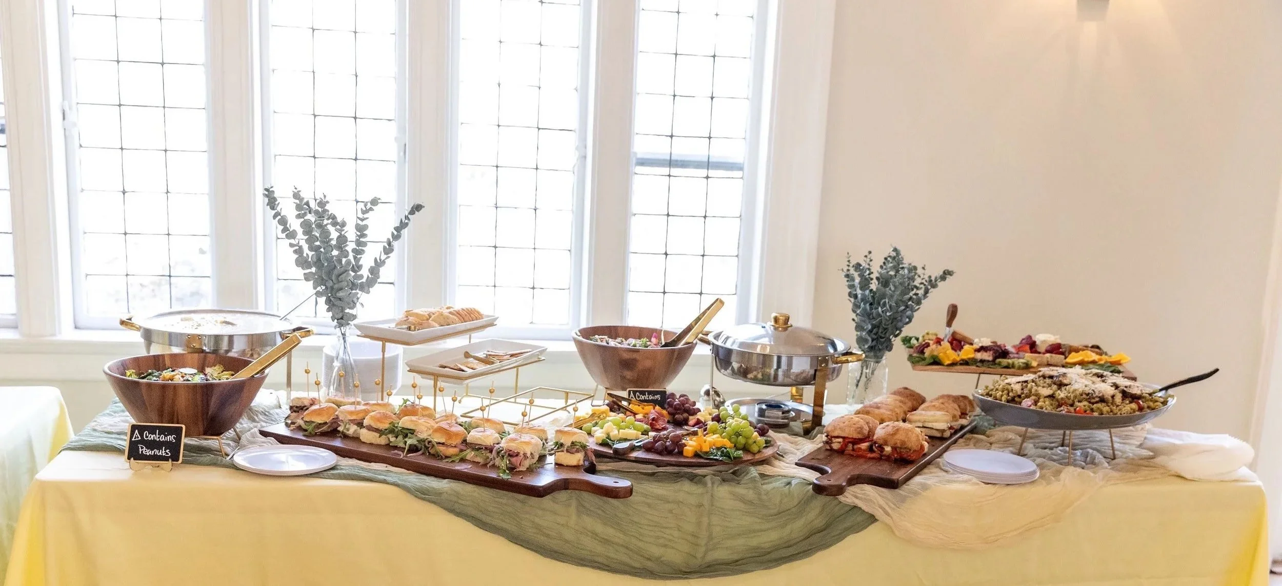 Buffet table with sandwiches, fruits, salads, and snacks in a bright room with large windows.