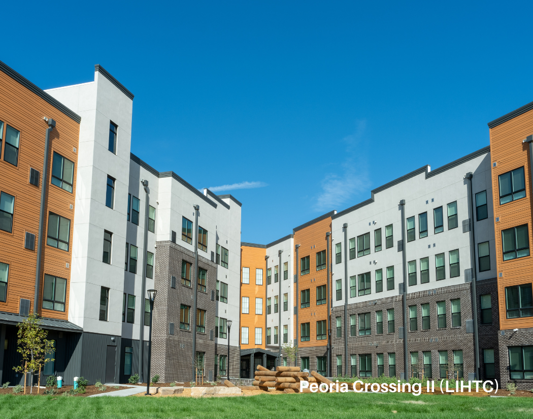 View of a modern apartment complex with multiple stories, white and brown facades, green lawn, trees, and a clear blue sky.
