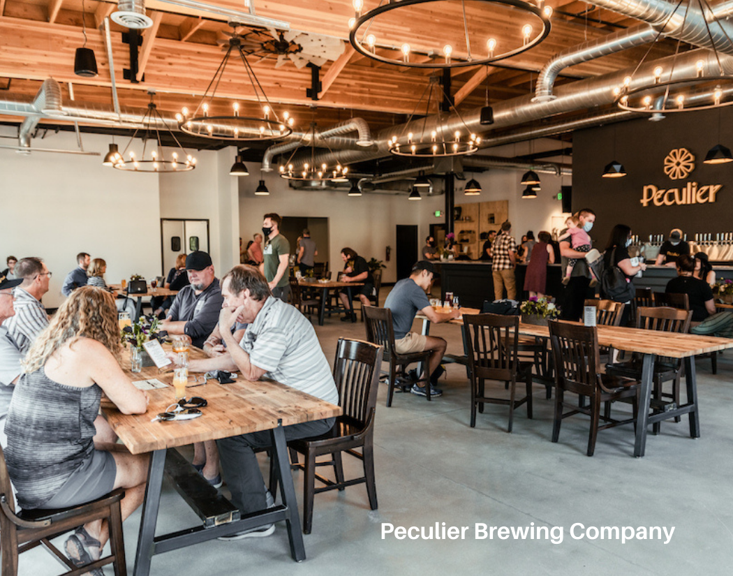 People sitting at tables and ordering drinks at Peculier Brewing Company, with industrial-style decor, including exposed ductwork and circular chandeliers.