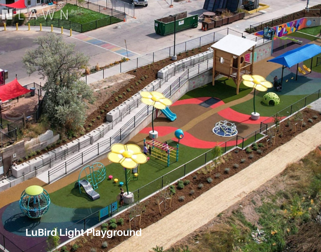 A colorful children's playground with slides, climbing structures, and seating areas, surrounded by a fence and landscaping.