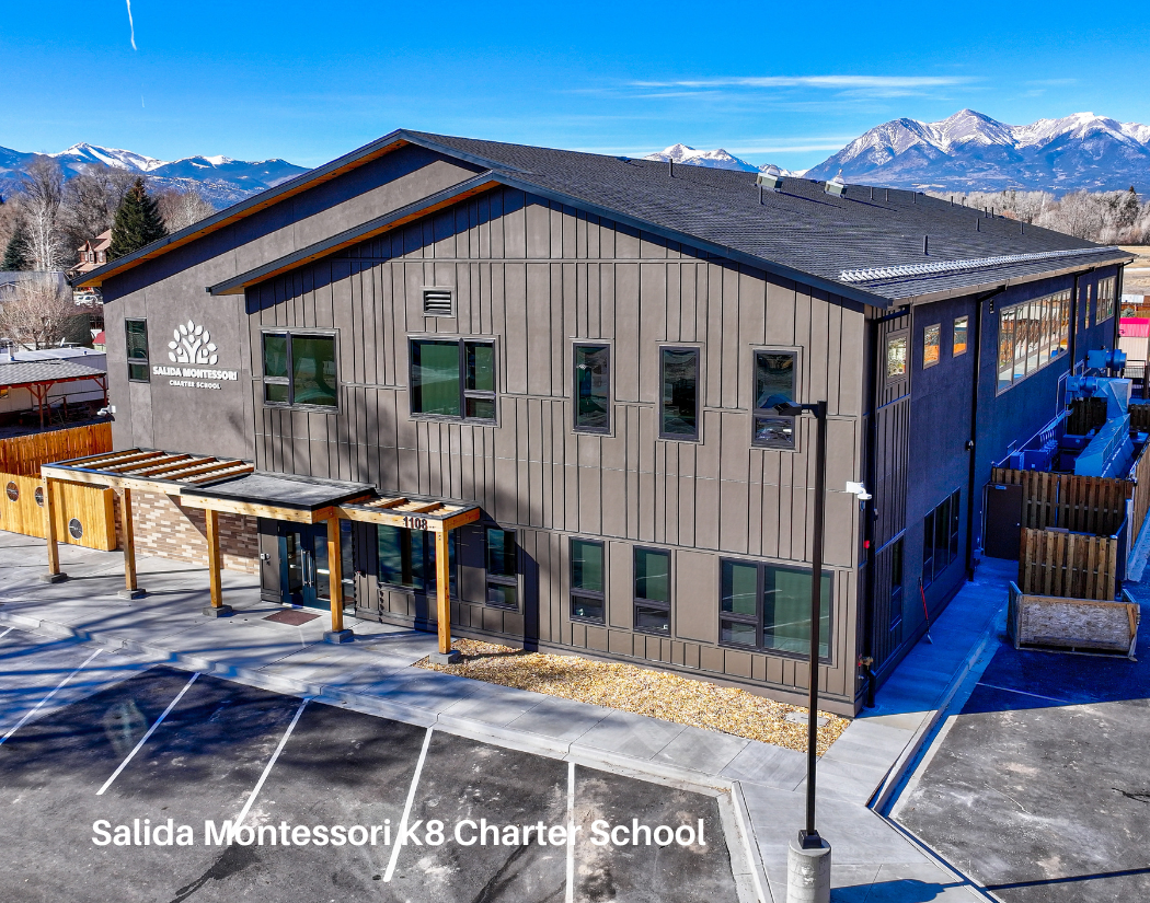 A modern, two-story school building with wooden and metal siding, labeled 'Salida Montessori K-8 Charter School,' with a parking lot in the foreground, surrounded by mountains and clear blue sky.