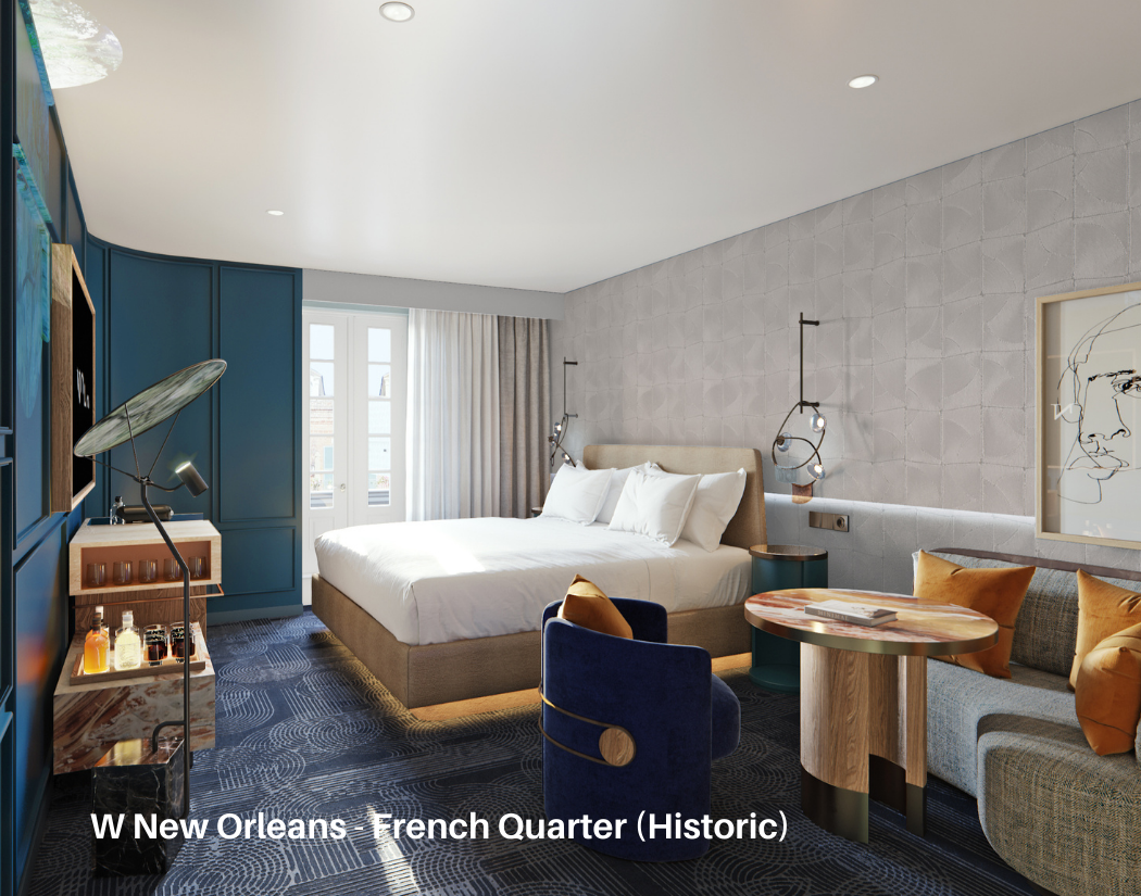 A modern hotel room with a large bed, blue accent wall, abstract artwork, round wooden table, blue and gray chairs, and natural light coming through French doors.