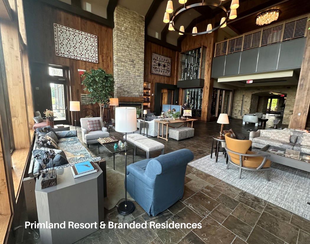 Interior of a hotel lobby with wooden walls, stone fireplace, and various seating areas including sofas, armchairs, and side tables with lamps. Large windows provide natural light, and decorative art pieces are displayed on the walls.