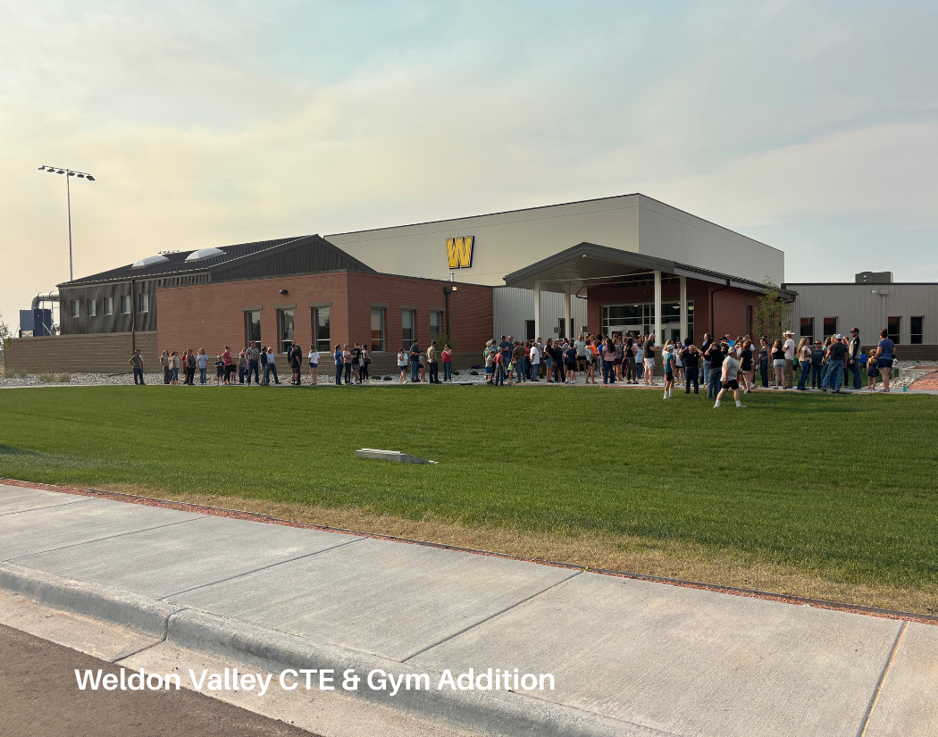 Long line of people waiting outside of a newly constructed building with a large yellow 'W' logo, labeled 'Weldon Valley CTE & Gym Addition', on a bright, partly cloudy day.