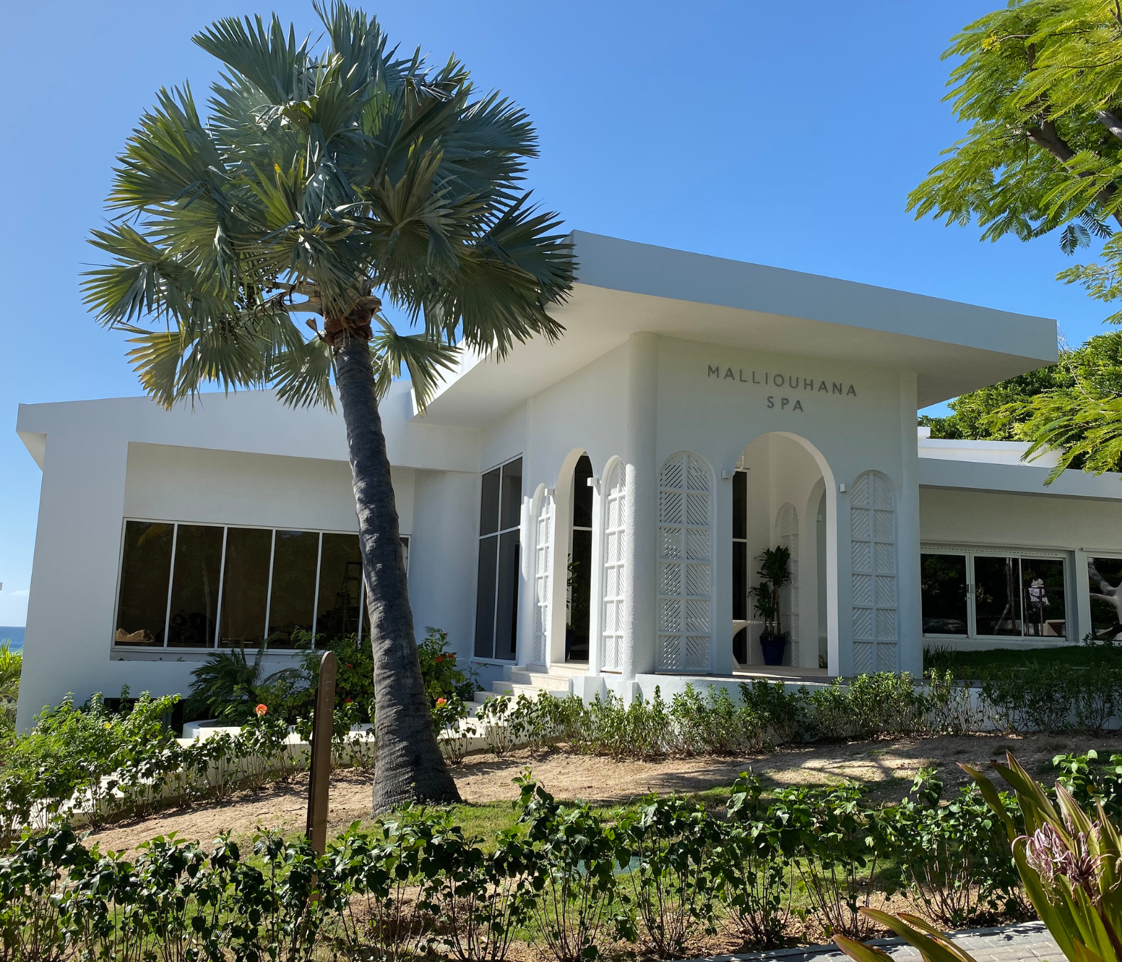 White building labeled "Malliouhana Spa" with palm trees and greenery in front, under a clear blue sky.