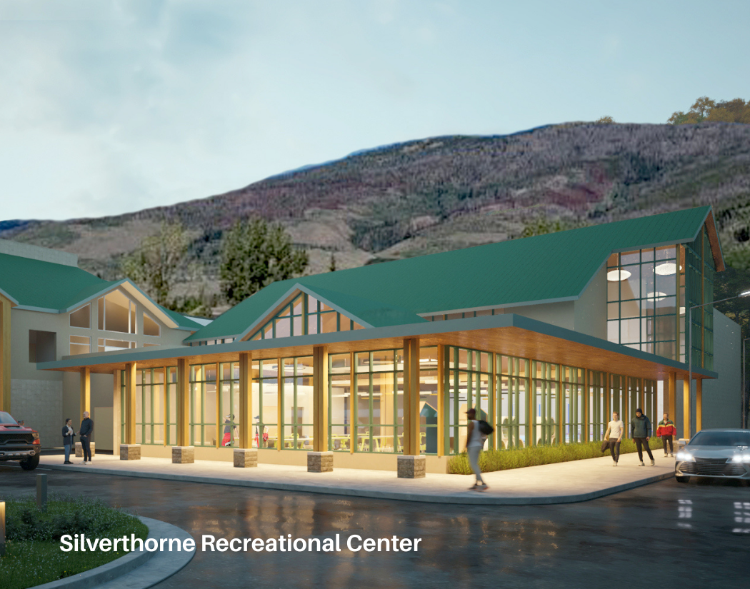 A modern recreational center with large glass windows, wooden accents, and a green metal roof, situated in a mountainous area, with people walking and cars parked outside.