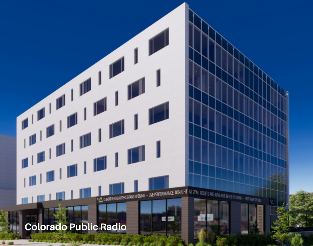 Modern multi-story building with a white facade and large windows, featuring a digital news ticker on the ground level, with trees and a clear blue sky in the background.