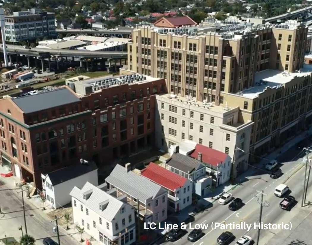 Aerial view of a city street with large historic buildings, smaller houses with colorful roofs, parked cars, and a highway in the background, during daytime.