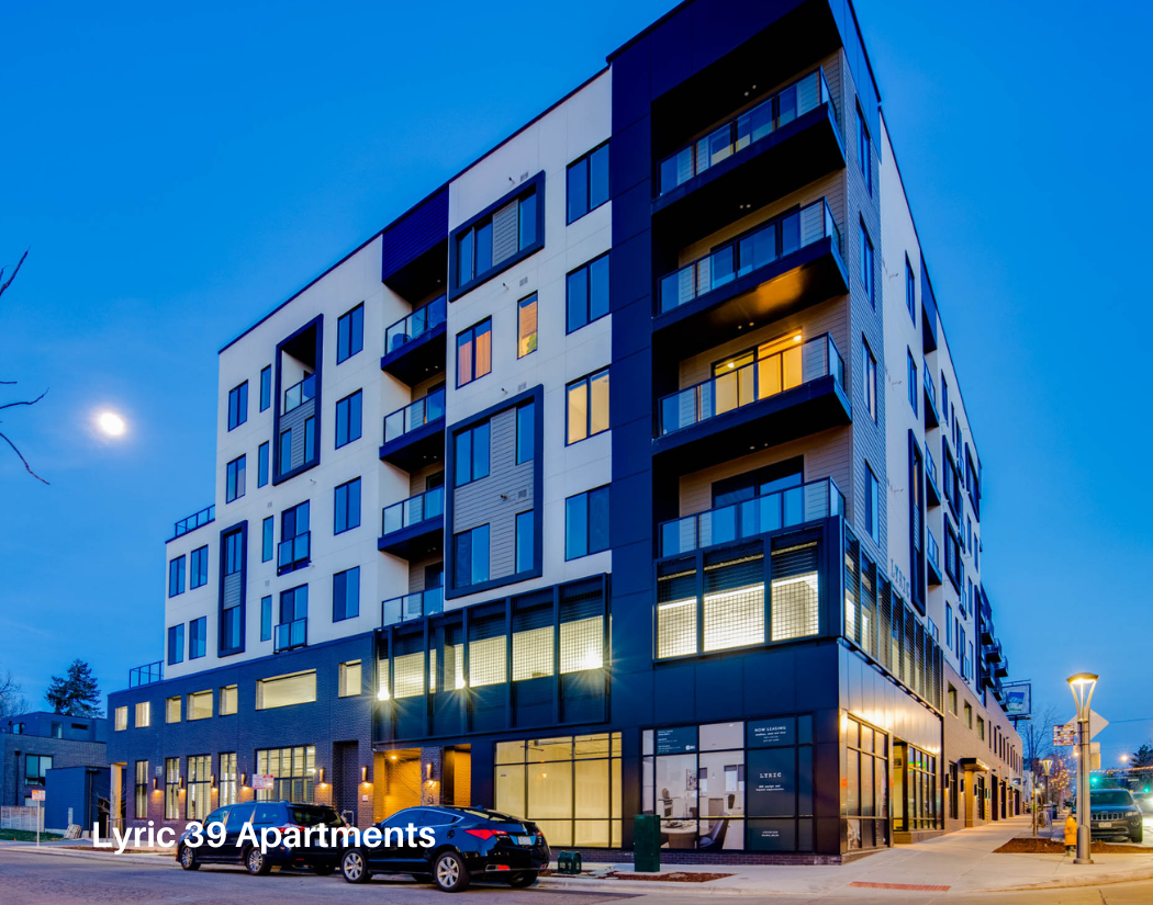 Night view of Lyric 39 Apartments, a modern multi-story residential building with a mix of glass and panel exterior, balconies, and illuminated ground floor with parked cars in front.