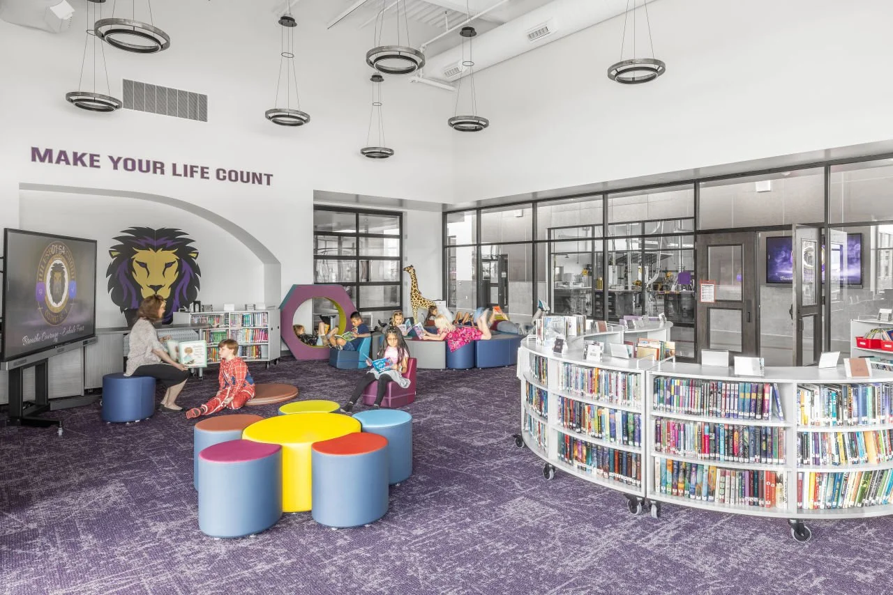 Children and a woman in a library children's reading area. Bright space with bookshelves, colorful furniture, and a giraffe statue. Sign on the wall says "Make Your Life Count."