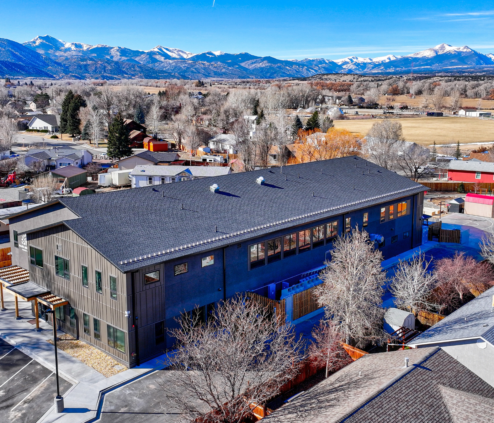 Aerial view of a modern building with a dark roof in a residential area, snow-covered trees, and mountains in the background.