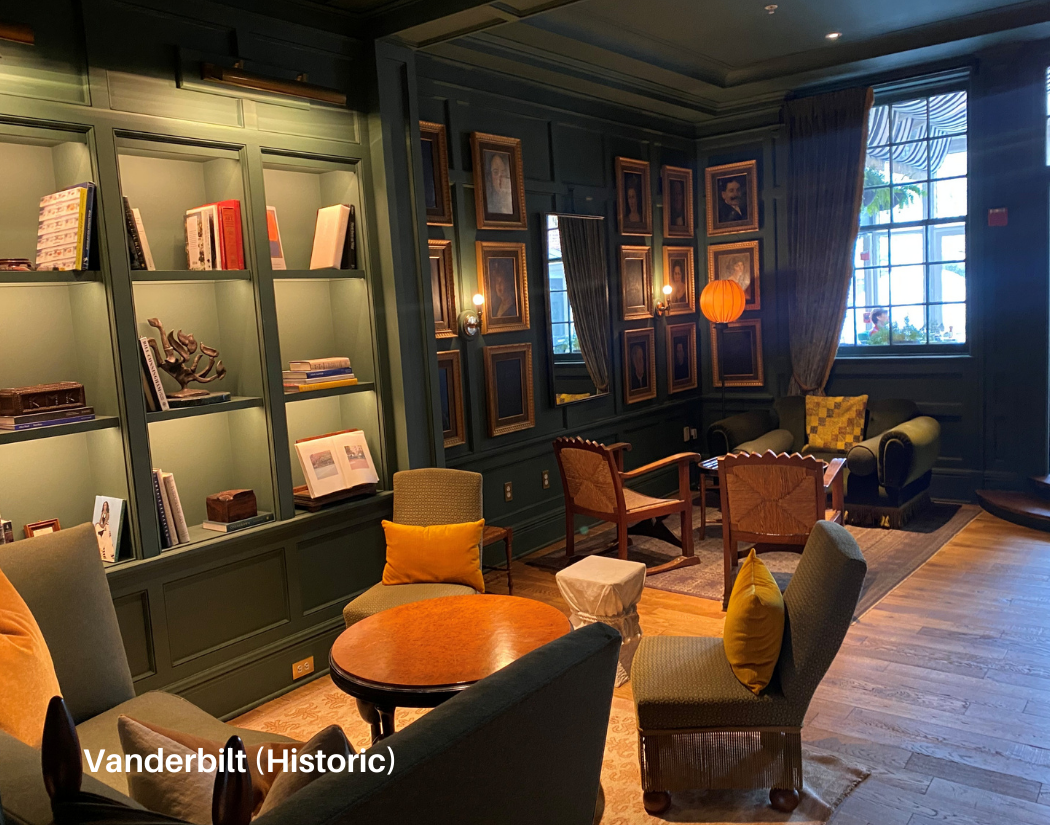 A cozy, vintage-style lounge or sitting room with dark green walls, picture frames, and bookshelves. There are several upholstered chairs with pillows, a small round wooden table, a sofa, large windows with curtains, and warm lighting including a han