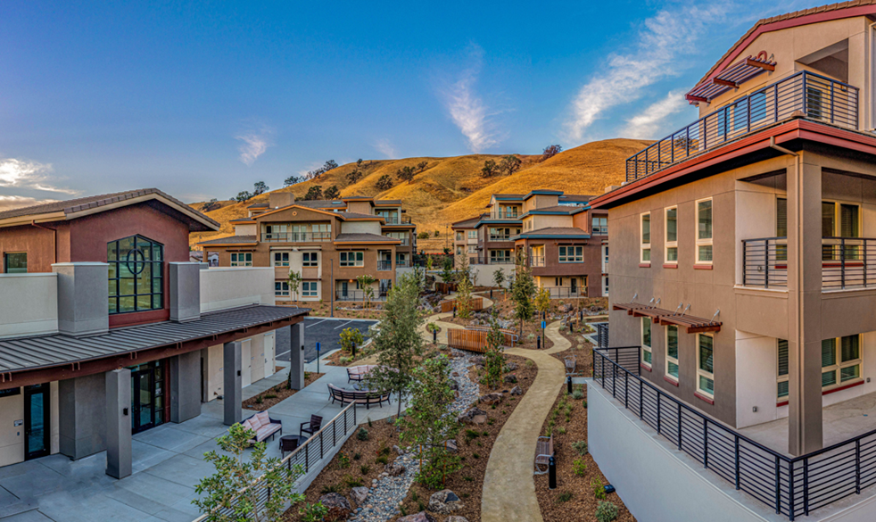 View of a modern residential complex with multiple apartment buildings, landscaped pathways, and a hilly background during daytime.