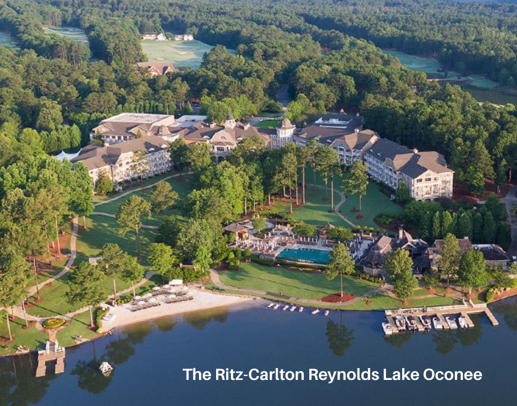 Aerial view of The Ritz-Carlton Reynolds Lake Oconee, showcasing a large resort complex surrounded by lush green trees, a lake with a beach and boats, swimming pools, and walking paths.