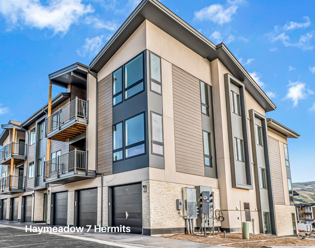 Modern multi-story residential building with garages and balconies, situated in a sunny, open area with blue sky and hills in the background.