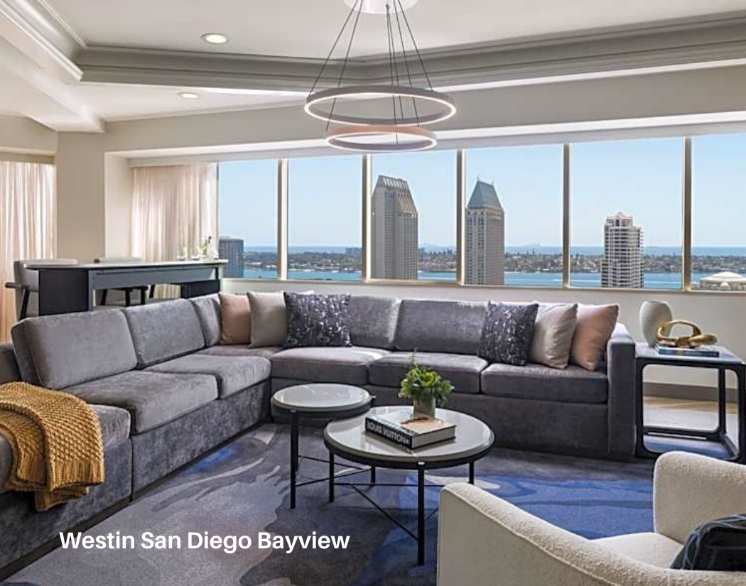Living room with large window view of cityscape and water, gray sectional sofa with pillows, modern light fixture, coffee tables with books and plant, beige armchair, side table with decorative objects.