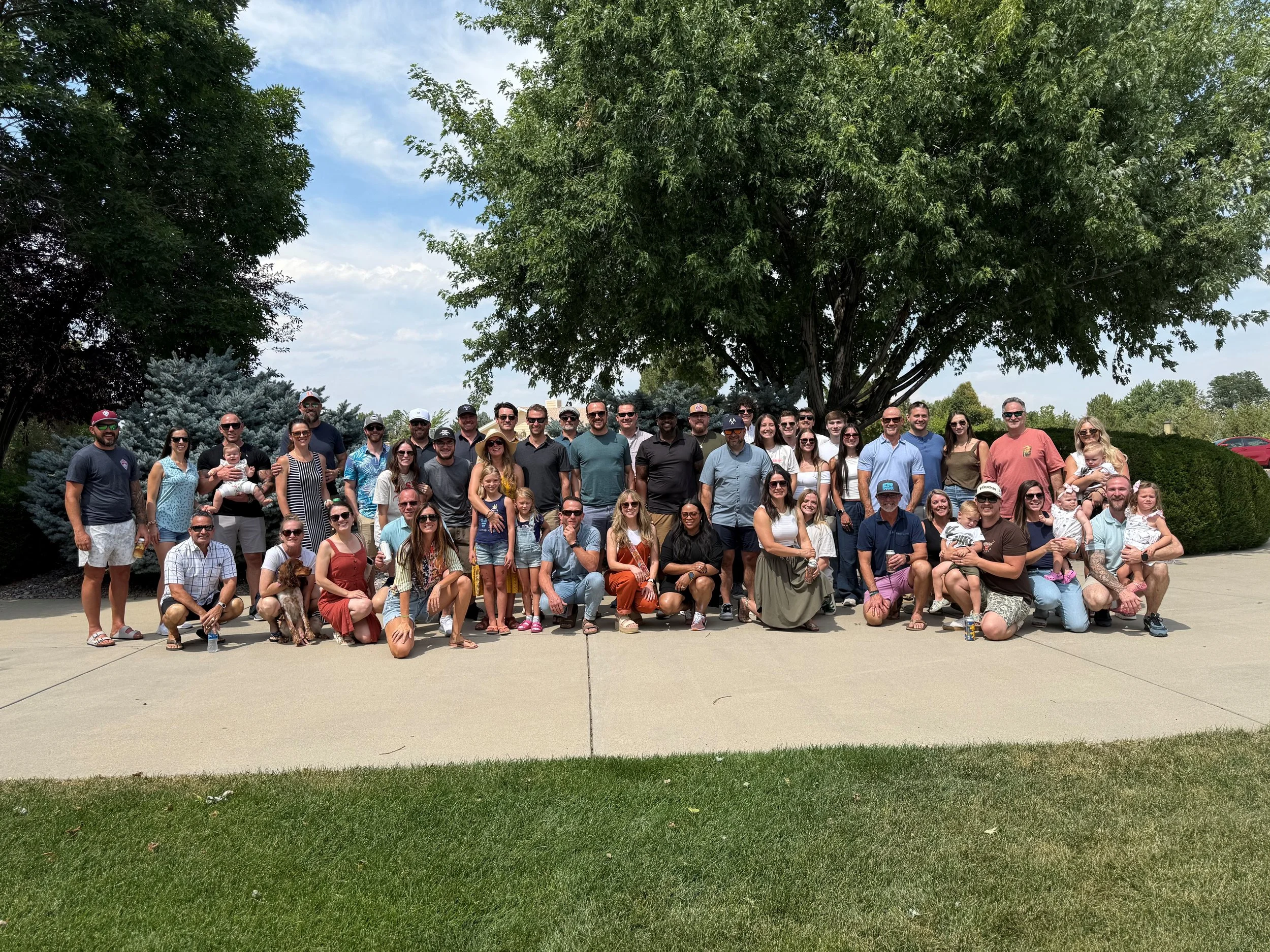 A large group of people posing for a photo outdoors on a sidewalk, with a big tree and blue sky in the background. The group includes adults, children, and a dog.