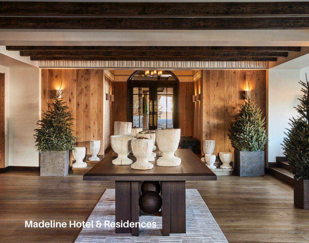 A hotel lobby with a dark wood table in the center, decorated with abstract white sculptures. There are potted Christmas trees on each side of the room, with wooden wall paneling and ceiling beams. Soft lighting is on the walls and a glass door at th