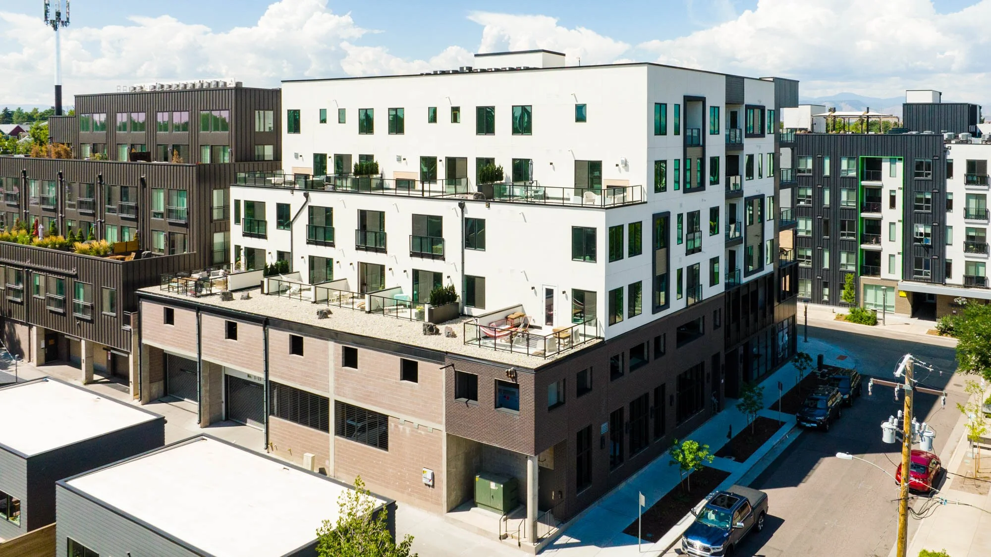 A modern multi-story apartment building with rooftop patios, large windows, and a street with parked cars and utility poles in a sunny urban area.