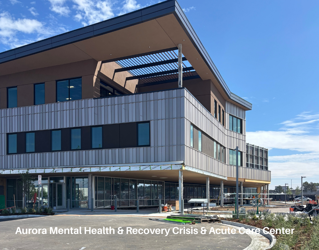 Under construction modern building with multiple floors, large windows, and outdoor terraces, labeled Aurora Mental Health & Recovery Crisis & Acute Care Center, on a sunny day with a blue sky.