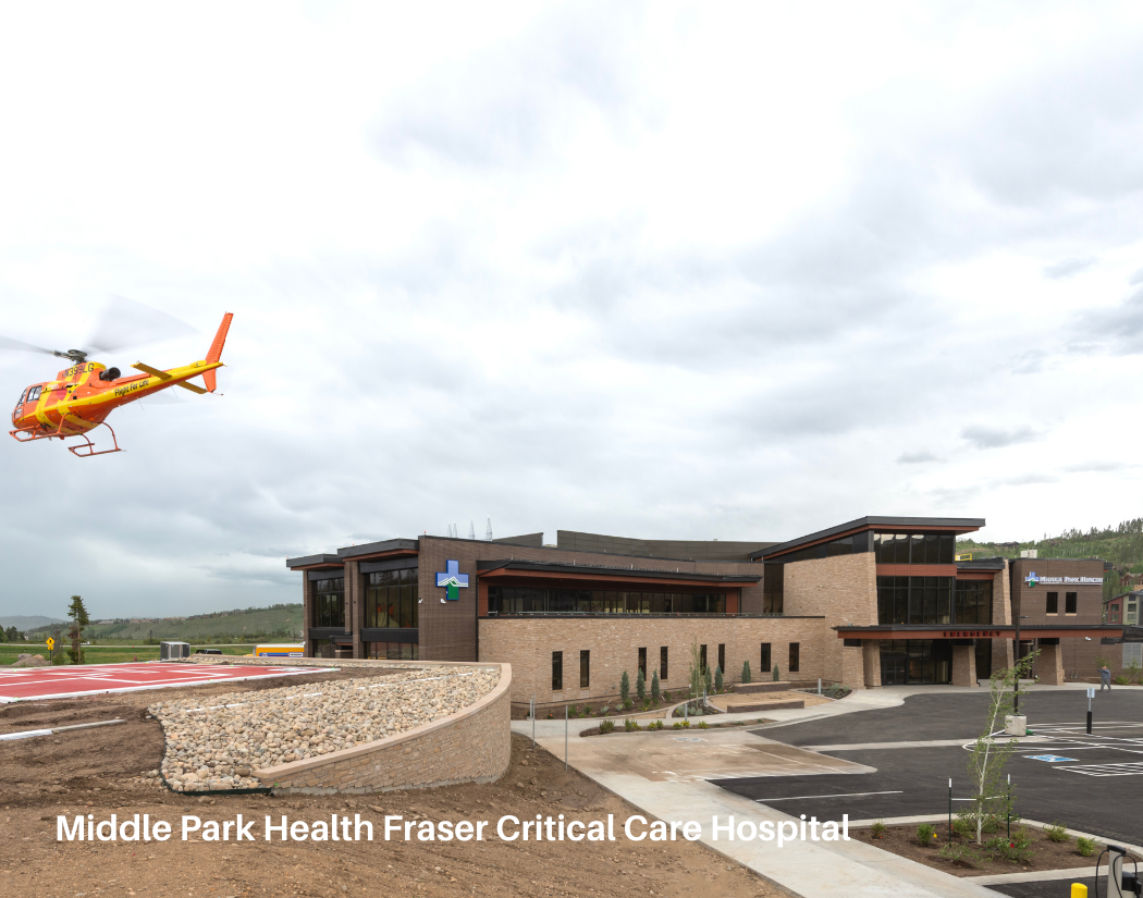 A hospital building with a parking lot and a plastic cross sign, with a yellow and red helicopter flying in the cloudy sky.
